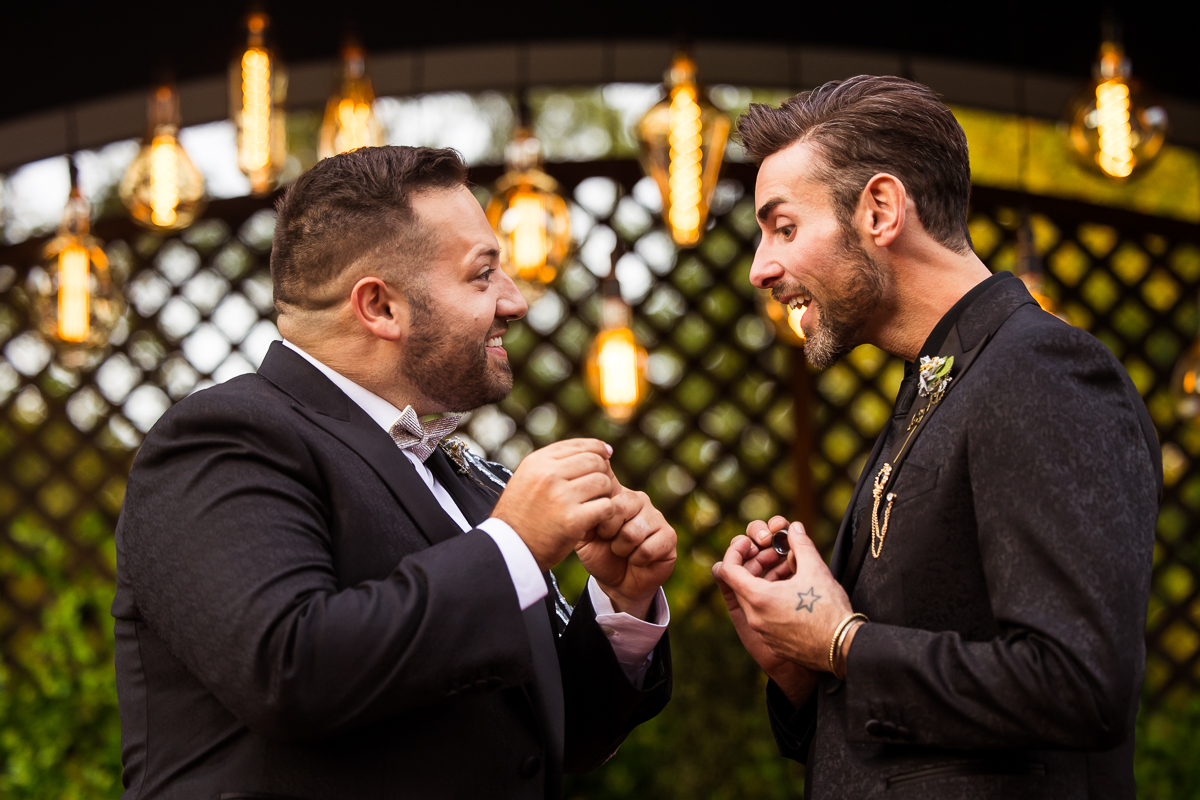image of the groom giving the other groom surprising him with a new ring in the middle of their wedding ceremony with unique lights hanging in the background behind them 