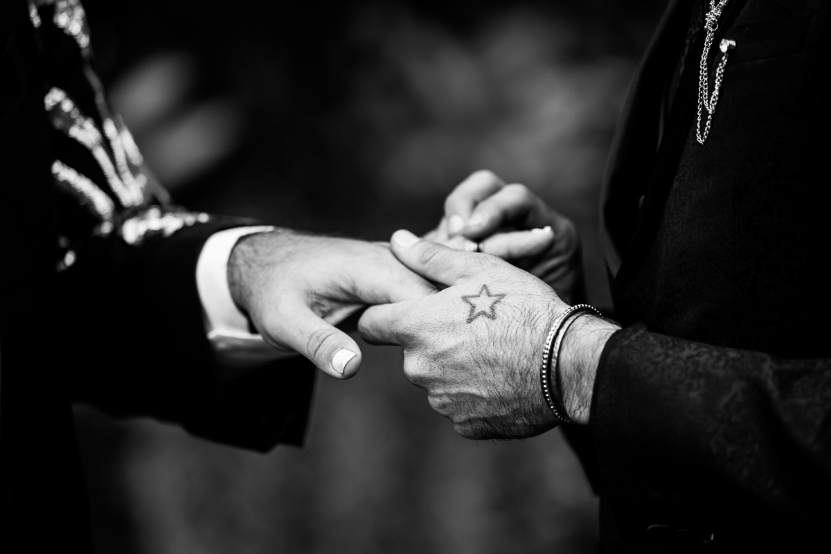 black and white close up image of the couple exchanging rings with one of the grooms having a star tattoo on their hand 