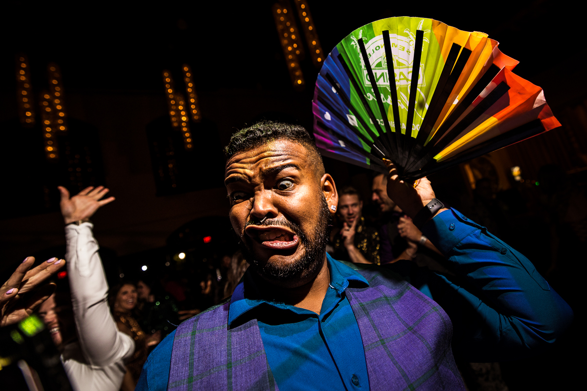 image of a guest making a funny face as he holds a rainbow fan up behind their head during this LGBT wedding reception 
