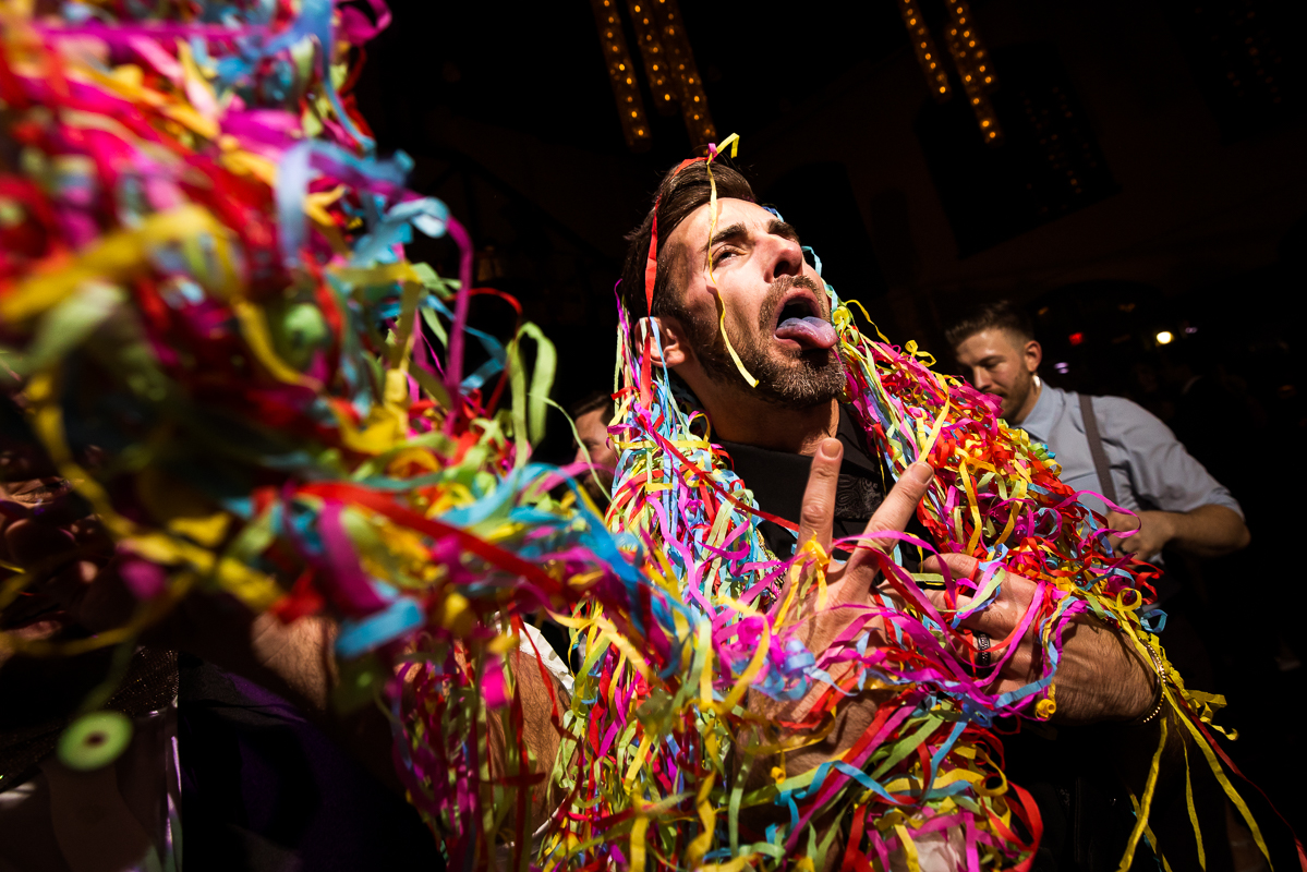 colorful, vibrant, creative image of the groom sticking his tongue out as he is covered in all of the colorful streamers