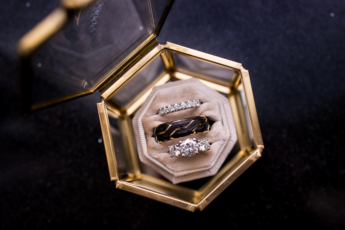 Image of the shiny diamond and black and gold rings in a glass and gold box ontop of a blue background during detail photos at this ritz carlton wedding ceremony in Washington dc