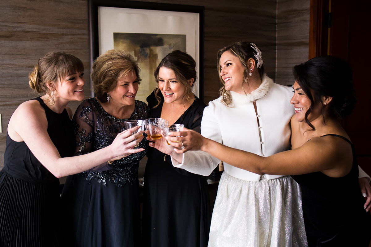Traditional image of the bride and her bridesmaids and mom holding drinks in together and saying cheers before the wedding ceremony 