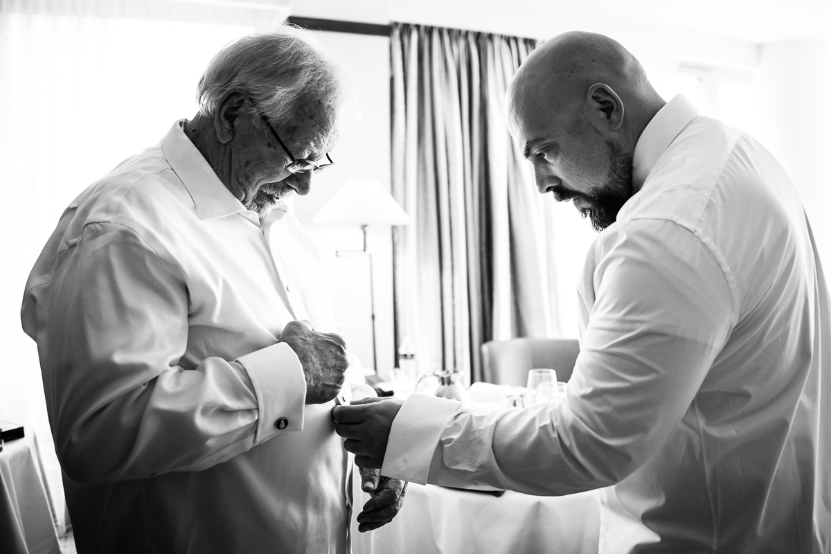 Black and white image of the groom helping his dad get ready and button his shirt during the preparations before their Georgetown wedding ceremony