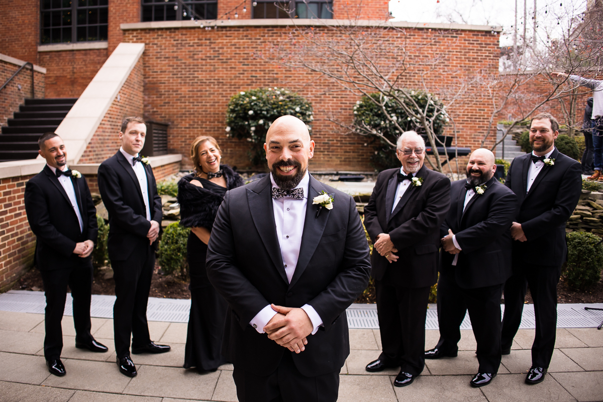 Traditional portrait of the groom and his groomsmen/women standing behind him in a straight line smiling at the camera with the red brick walls of the hotel behind him and a little bit of green shrubbery before their ritz carlton wedding ceremony 