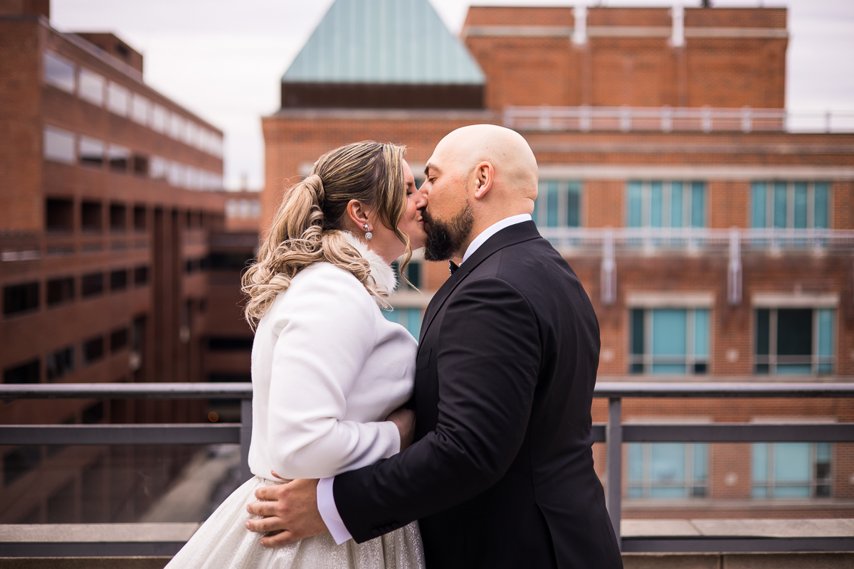Ritz Carlton Georgetown Wedding photographers, rhinehart phtoography, captures the couple as they kiss on the balcony outside of the hotel during their first look with red brick building filling the background behind them 
