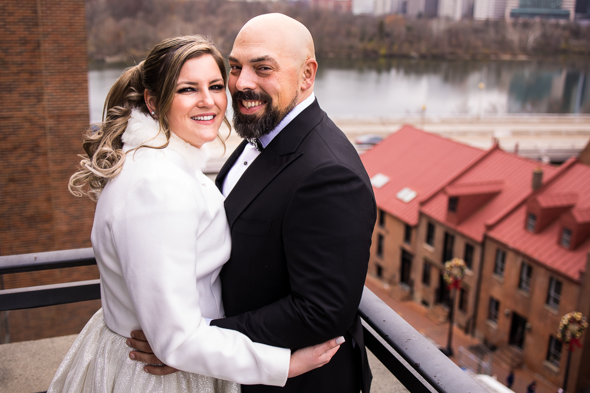 Traditional portrait of the couple hugging one another and smiling at the camera as there are historic buildings behind them as well as the Georgetown waterfront in the background as they take their romantic portraits during the Washington dc wedding 