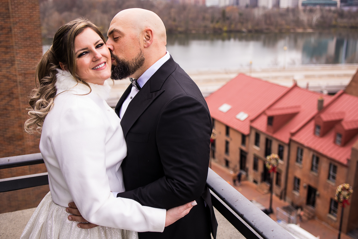 Traditional portrait of the couple hugging one another, kissing and smiling at the camera as there are historic buildings behind them as well as the Georgetown waterfront in the background as they take their romantic portraits during the Washington dc wedding 