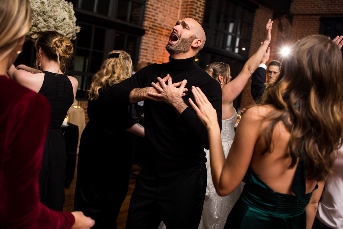 image of guests dancing and singing during this indoor wedding reception as one of the guests is singing his heart out with his hands on the chest 