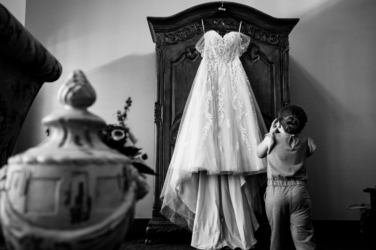 wedding photographer, lisa rhinehart, captures this black and white image of the brides dress hanging off a dresser as a little girl is standing near it looking at it before the stroudsmoor wedding ceremony 