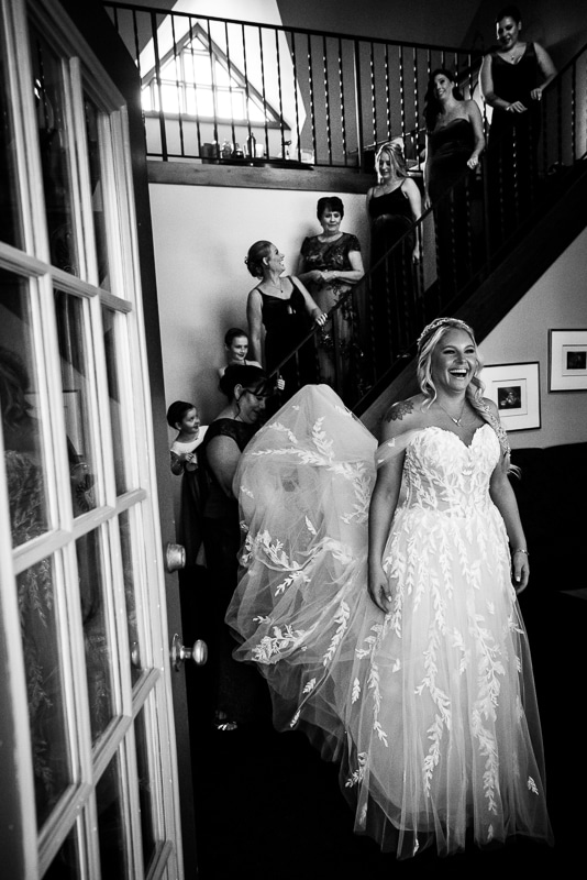 black and white image of the bride smiling as her bridesmaids line up on the stairs to see her and her dress for a first look before the wedding ceremony