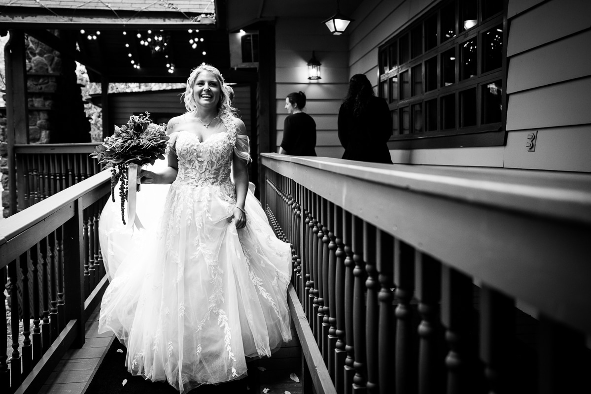 black and white image of the bride holding her dress and flowers as she walks across a bridge with a big smile on her face 