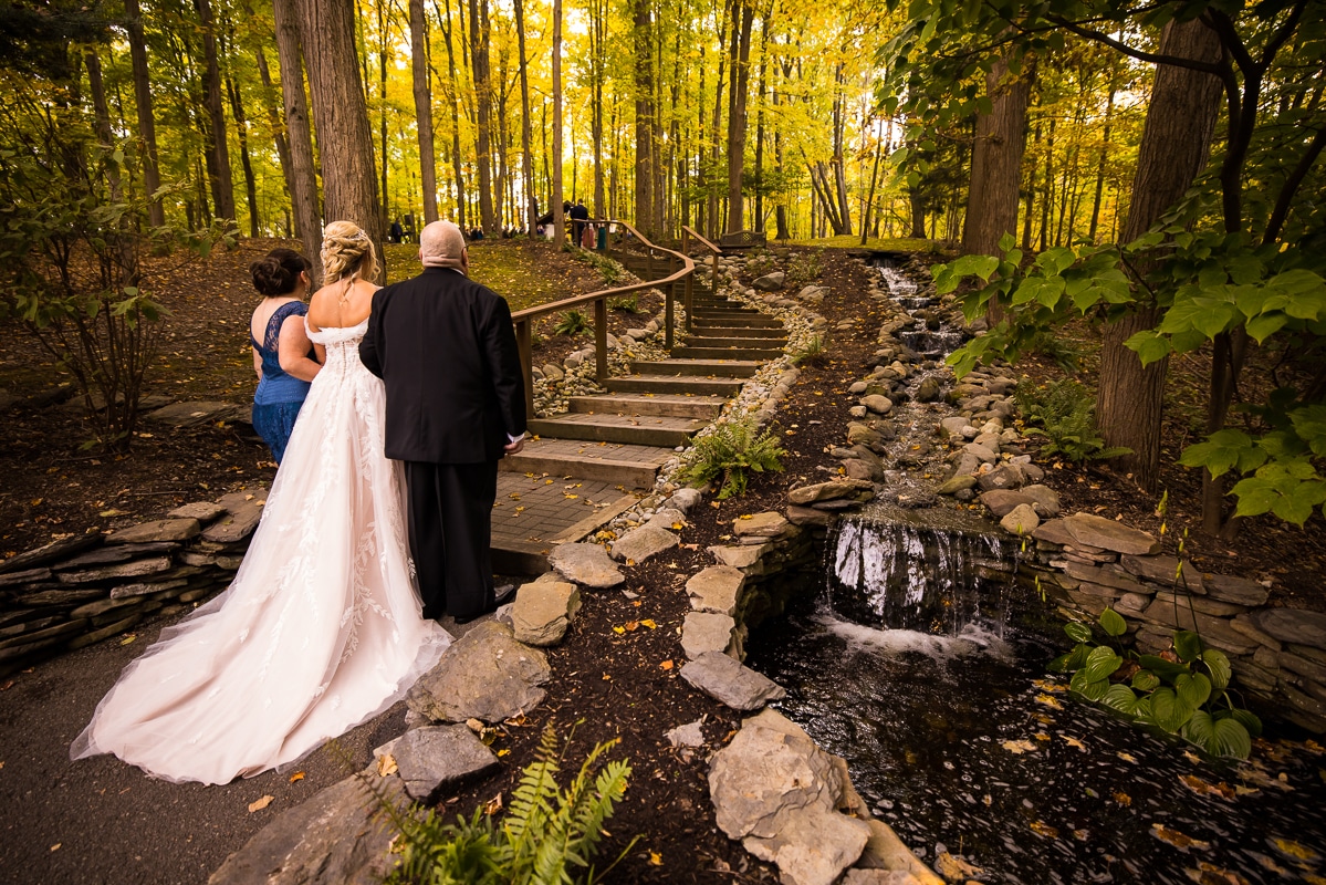 pocono wedding photographer, lisa rhinehart, captures the bride with her mom and dad as they walk up the walk way to the wedding ceremony surrounded by trees and a little waterfall at the stroudsmoor county inn woodsgate
