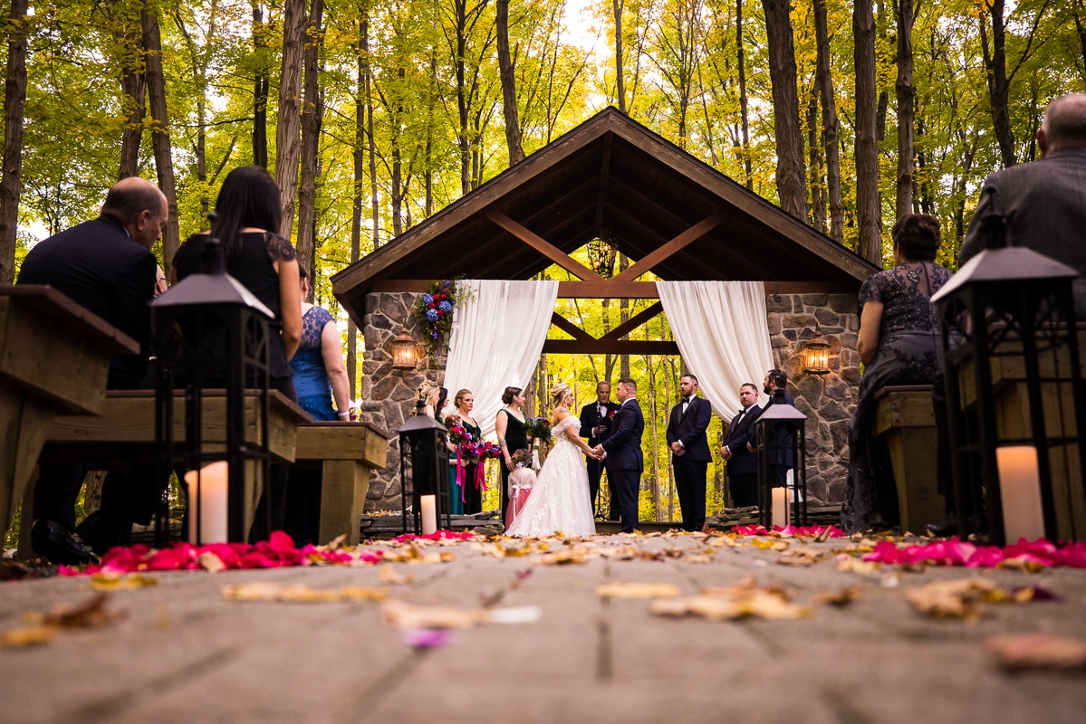 Pocono wedding photographer, lisa rhinehart, captures a unique and creative image of the couple as they stand under the wood and stone ceremony pavillion and captures the petals that have fallen on the stone walkway. you can also see the vibrant, colorful fall leaves that surround them on the trees at the stroudsmoor