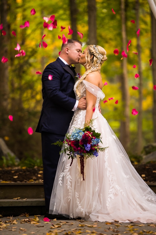 Pocono wedding photographer, lisa rhinehart, capture this unqiue, creative and playful image of the bride and groom kissing as bright pink flower petals blow around them in the wind as she holds her colorful flower bouquet in front of her dress