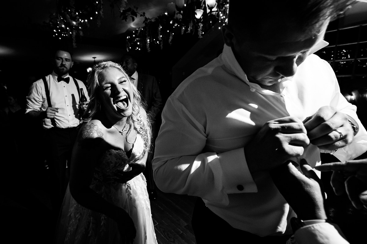 black and white image of the groom buttoning his shirt and the bride is standing behind him laughing and smiling as everyone dances together during their stroudsmoor wedding reception 