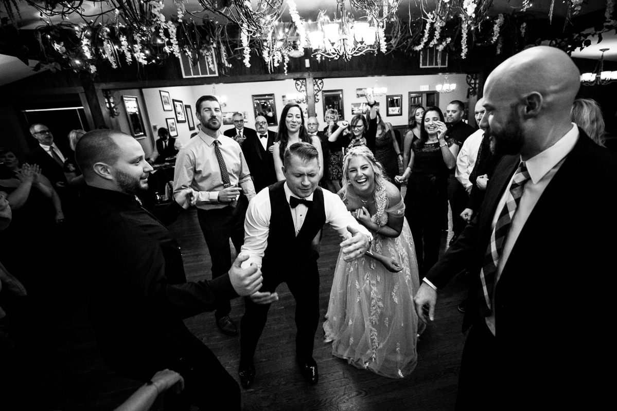 black and white image of the groom dancing and singing as the bride stands off to the side behind him laughing as he does this during their wedding reception