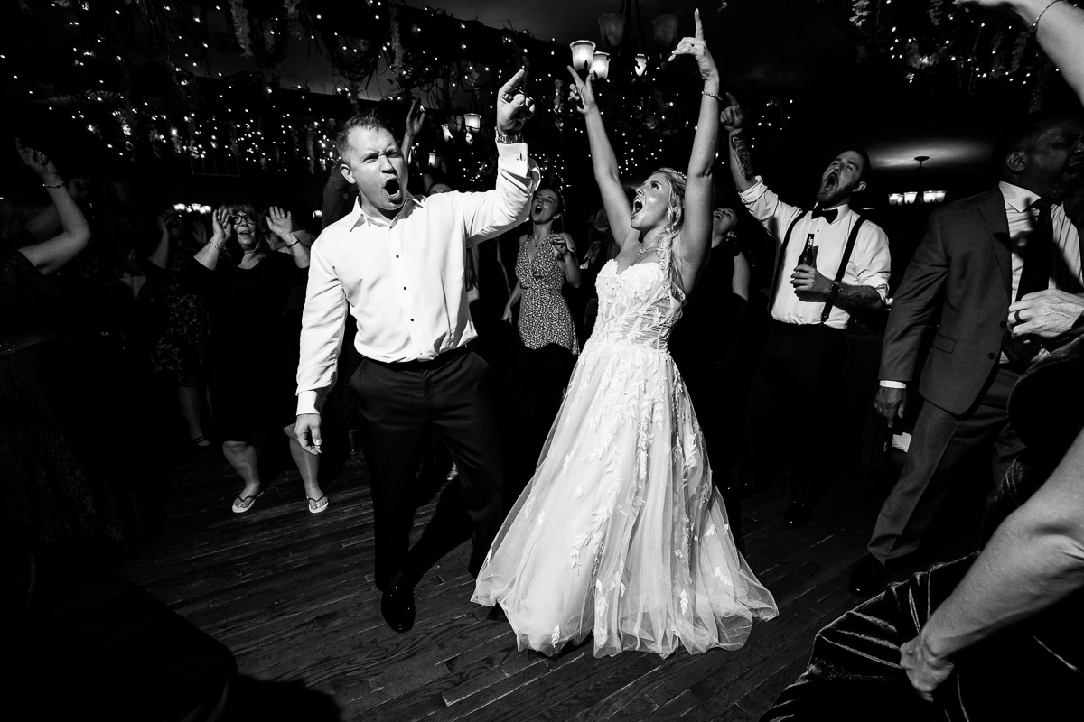 Pocono wedding photographer, lisa rhinehart, captures this black and white of the couple dancing with their friends and family as they have their arms up in the air with little lights filling the ceiling above them 