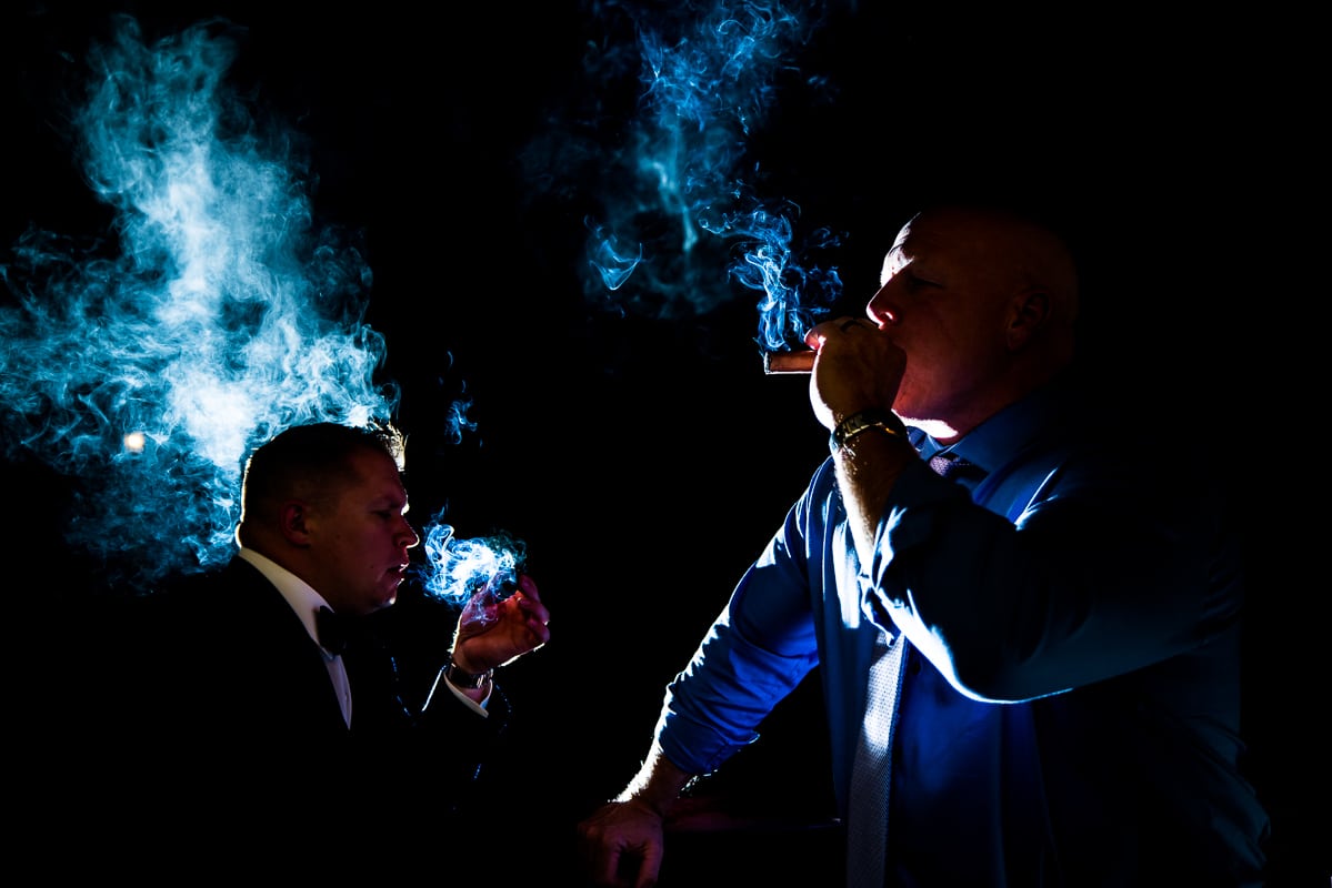 creative pocono wedding photographer, lisa rhinehart, captured this unique and creative image of the groom and friends smoking a cigar with the smoke blowing into the air with a blue tint to it from the back light 