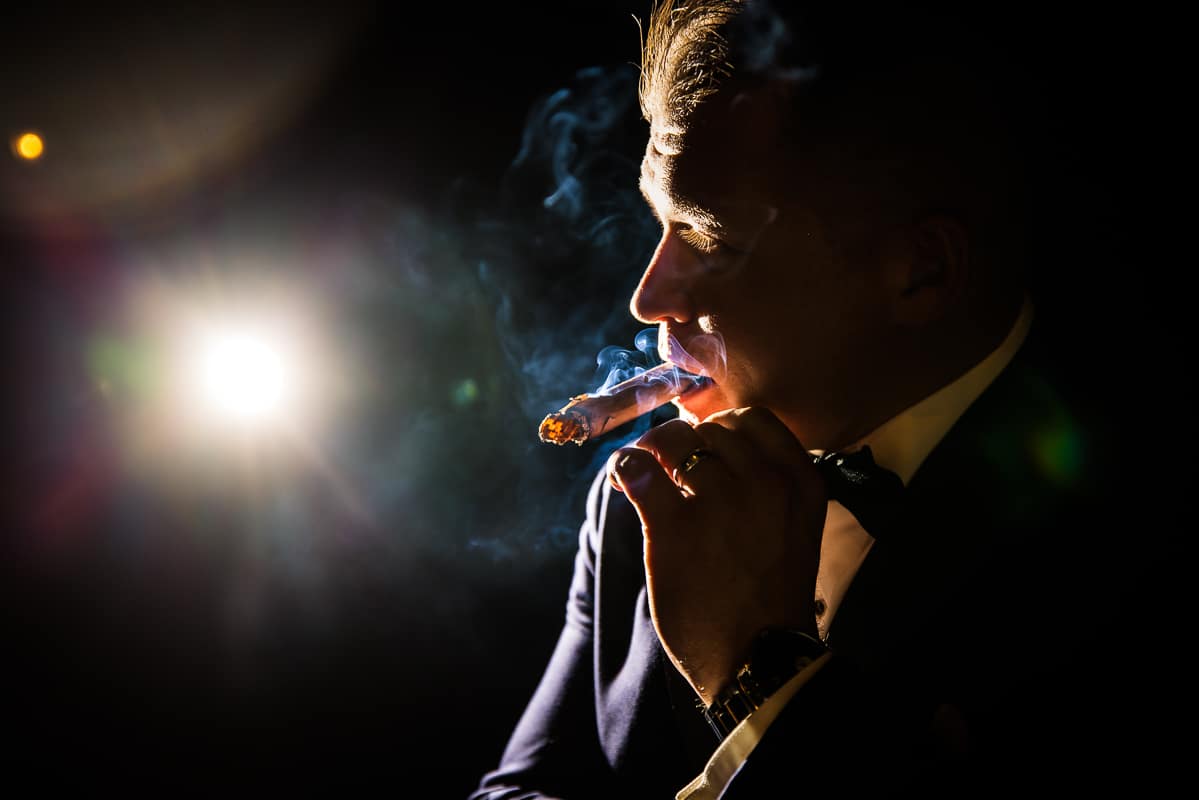 creative image of the groom smoking his cigar and the blue smoke rolling through the air as he is backlit outside of the stroudsmoor