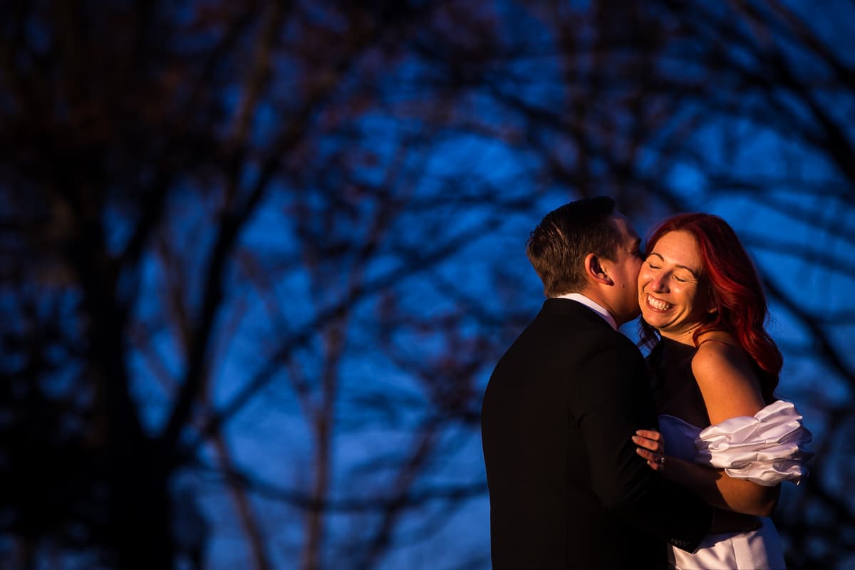 Vibrant, blue image of the couple hugging and kissing one another as there are tons of trees behind them at the mansion in Mechanicsburg, Pennsylvania