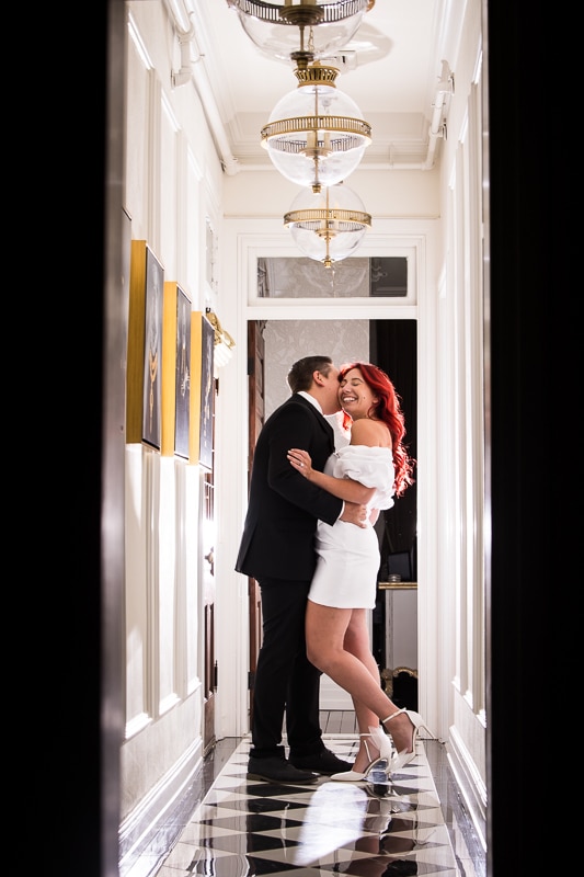 image of the couple hugging one another close as one of them kisses the other inside the mansion with checkered flooring and big circle chandeliers handing from the ceiling for this engagement session