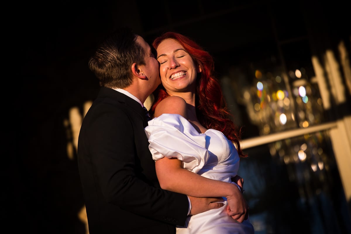 Image of the couples hugging from behind as they kiss on the cheek in their formal attire with glass reflections behind them during their Ashcombe Mansion Engagement