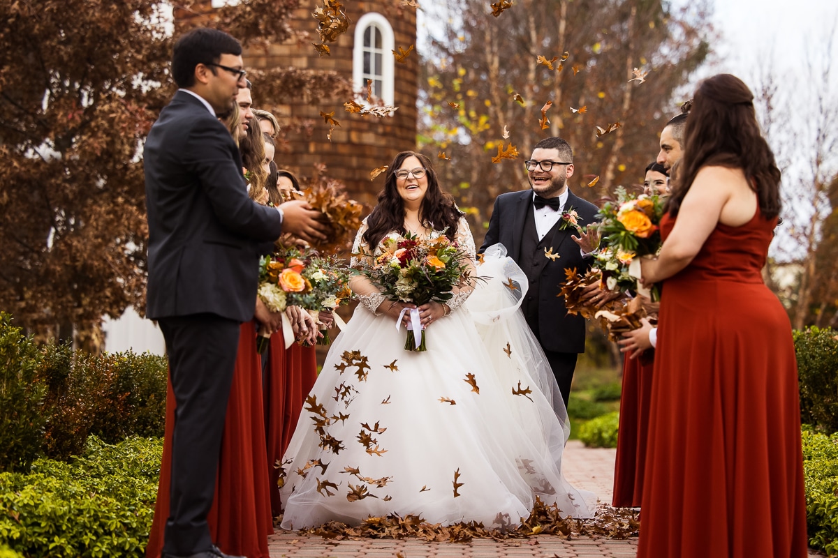 New Jersey wedding photographer, lisa rhinehart captures the couple laughing and smiling at their wedding party as they thrown leaves up in the air while the couple walks between them during their wedding day at the Ryland Inn, NJ