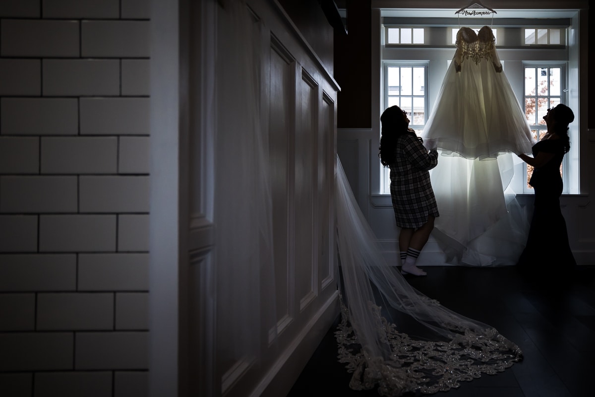 Unique, creative image of the brides dress hanging from above the window as her and her mom look up at it with the veil also in the picture on the left side 