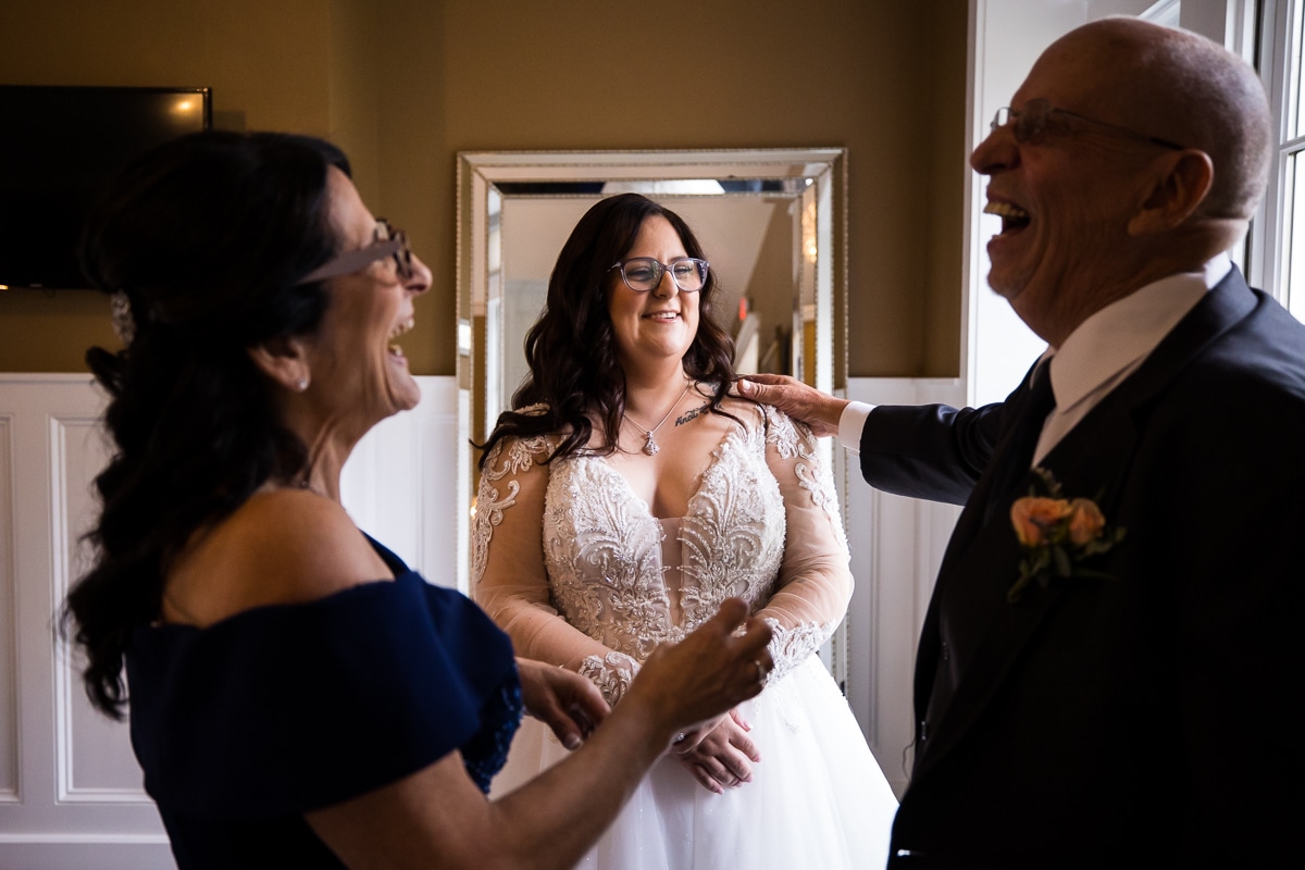 Image of the mom and dad laughing and smiling as the bride smiles at her dad during their first look before the wedding ceremony 