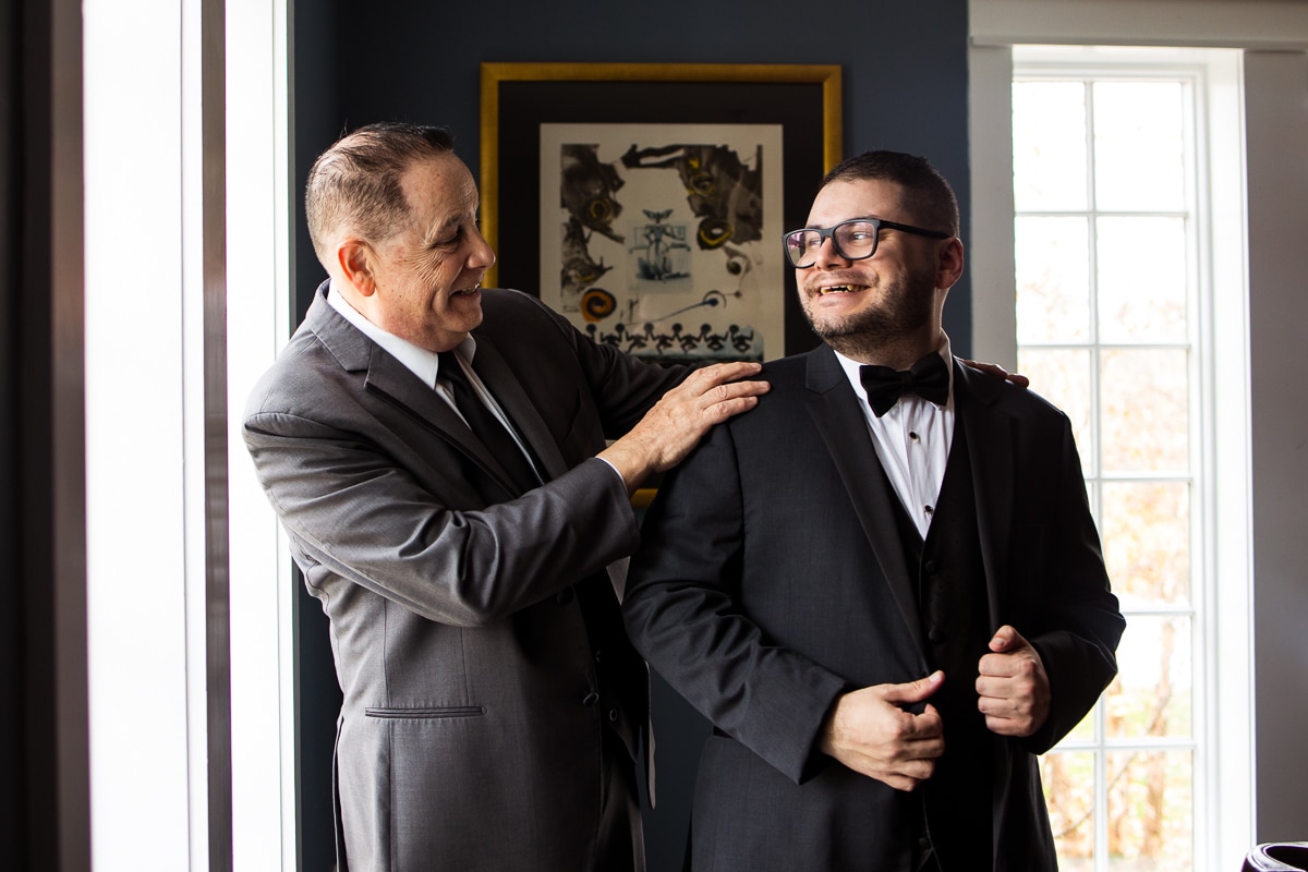 creative, candid image of the groom getting his tux on with the help of his dad as they smile at one another during the preparations before their wedding at the Ryland Inn 