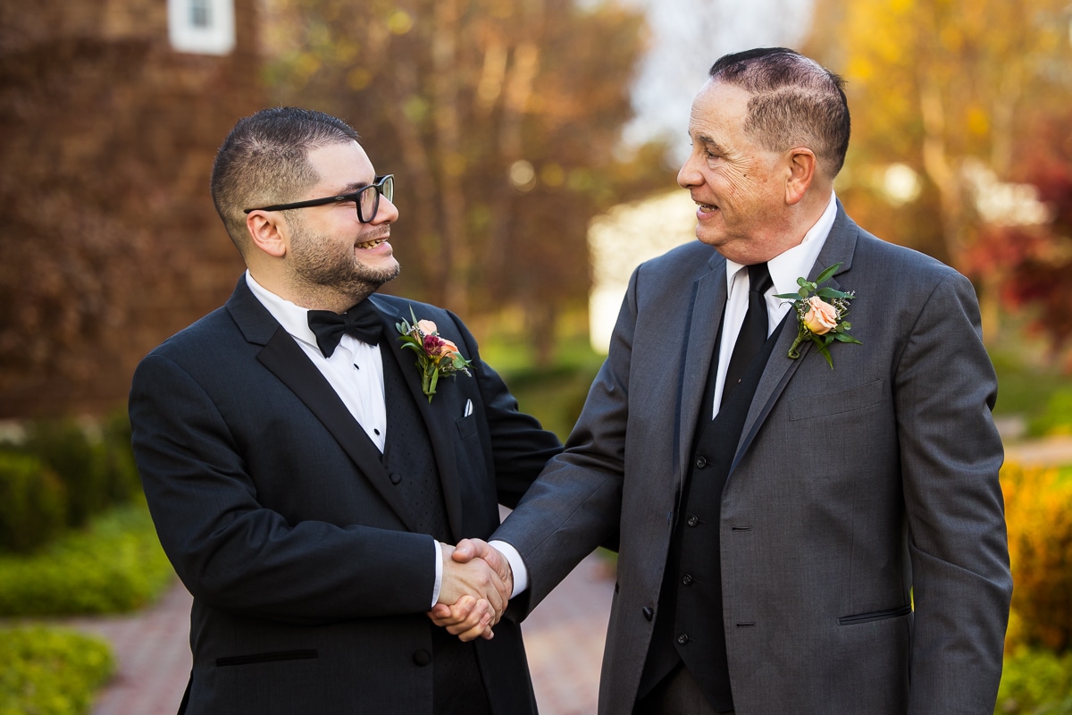 candid image of the groom holding his dads hand and shaking it as they smile at one another before the grooms wedding ceremony at the ryland inn 