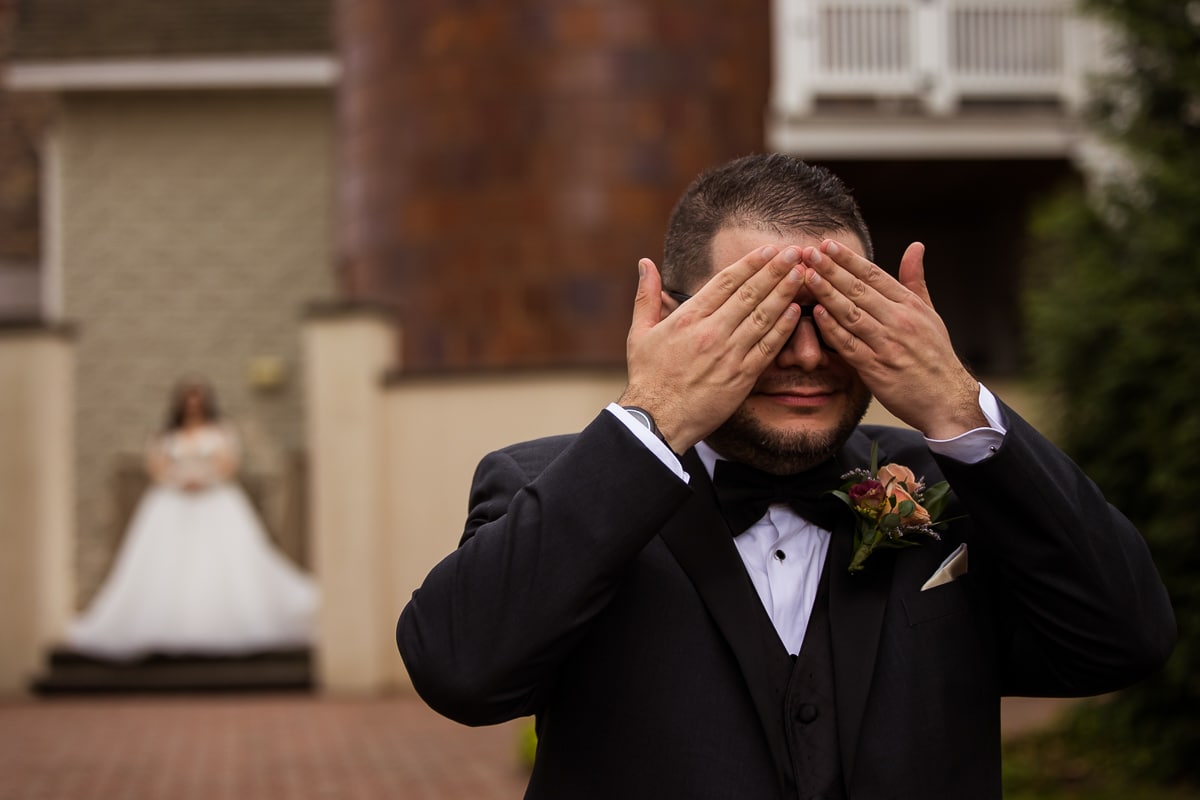 Close up image of the groom covering his eyes with the bride blurred out in the background as the couple prepares to have their first look outside of the Ryland Inn 