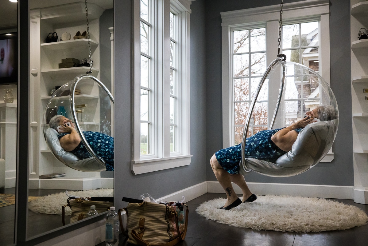 unique perspective of the grandma sitting in a bubble chair on the phone during wedding preparations and her reflection is captured in the mirror 