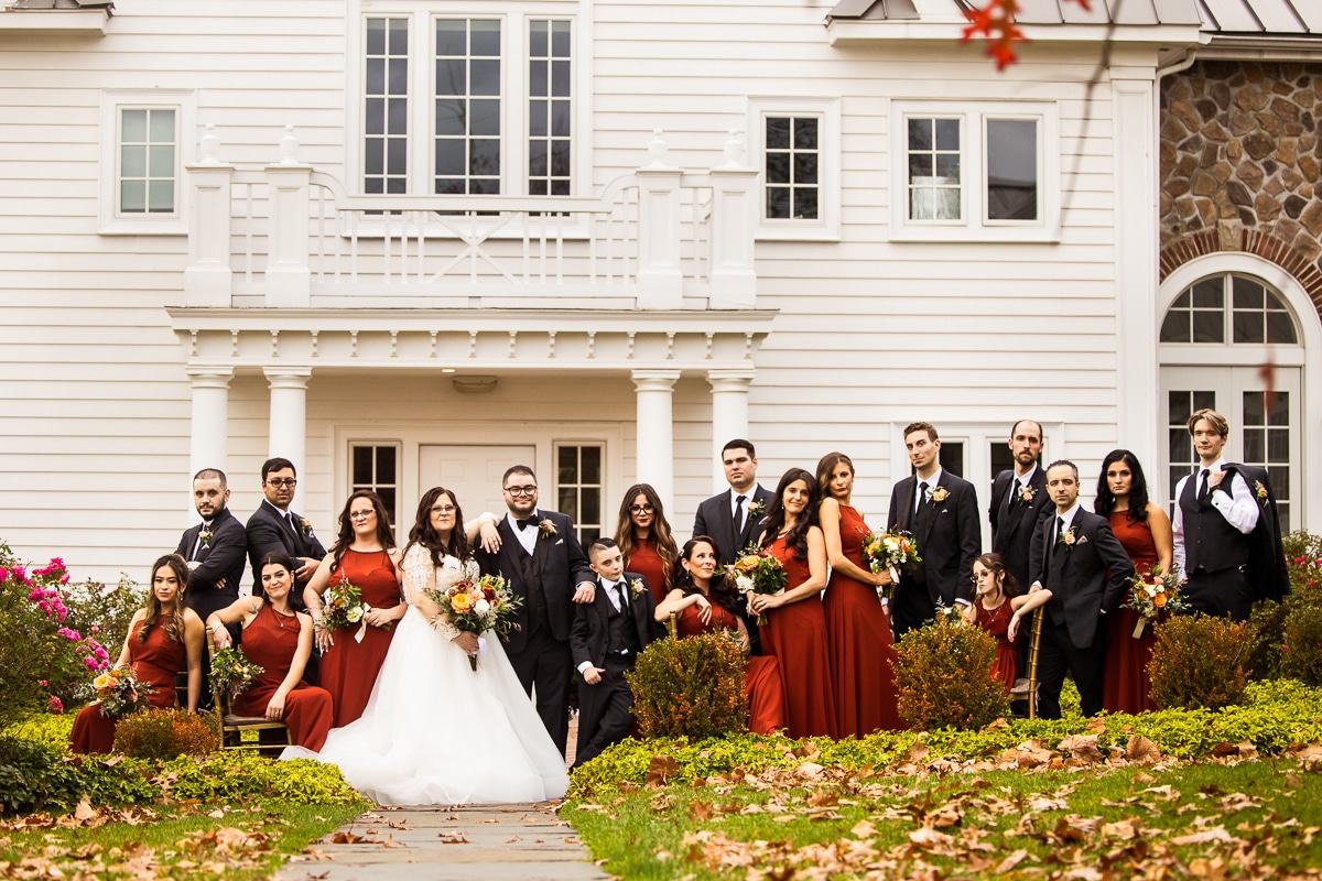 traditional image of the wedding party standing in different poses outside of the ryland inn before the wedding ceremony dressed in their black tuxedos and burnt orange/burgandy dresses