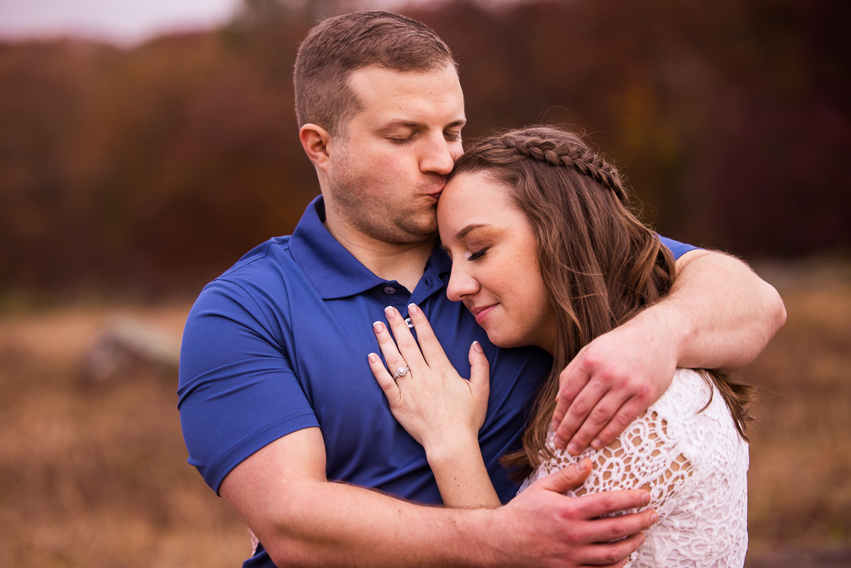 Traditional image of the couple hugging one another and kissing her on the forehead in their blue and white outfits during their outdoor, fall engagement sesssion