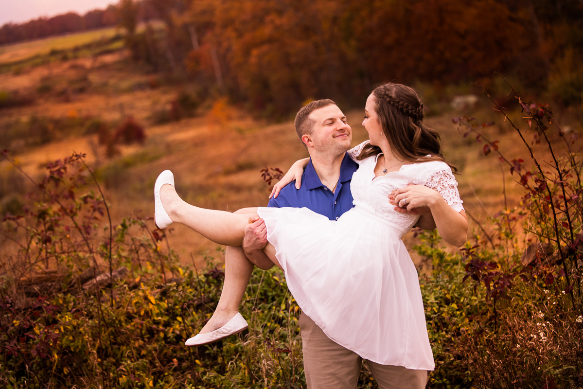 candid image of the guy picking up the girl in his arms and looking at her with a smile on his face surrounded by colorful fall foliage during their outdoor engagement session in Gettysburg