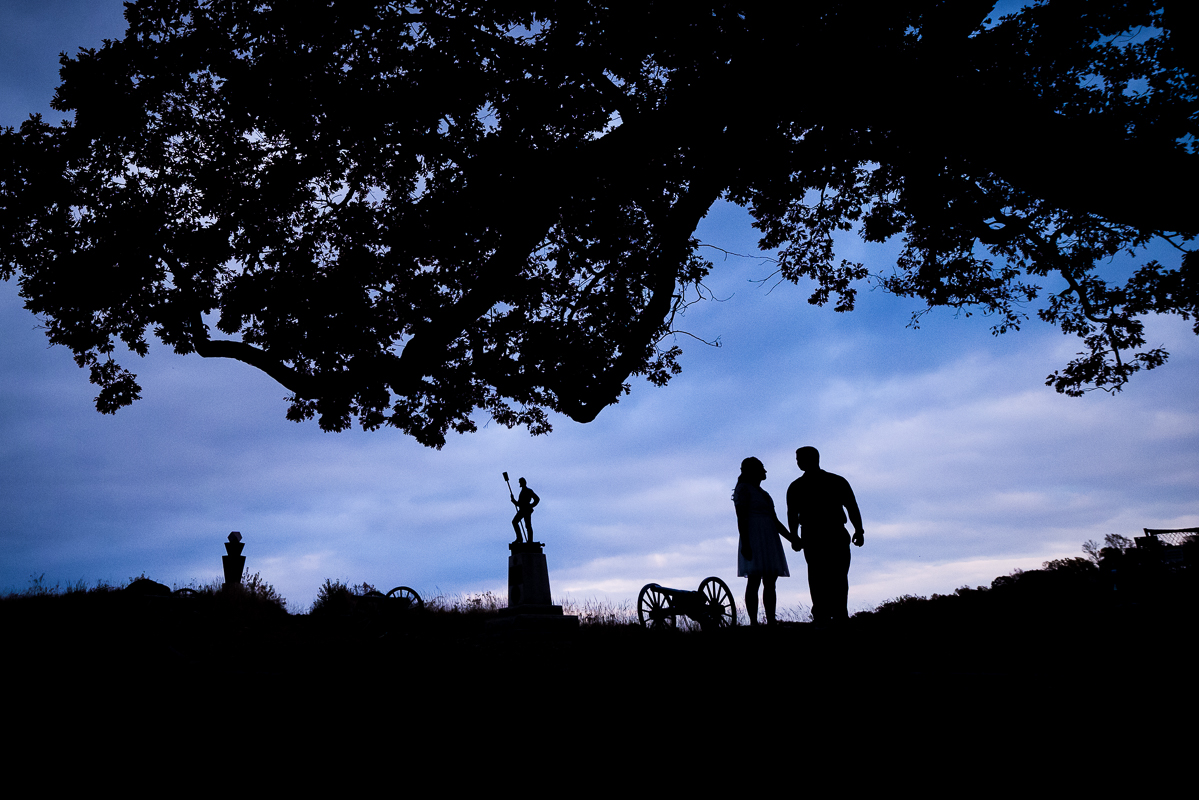 Devil's Den Gettysburg Engagement photographer, lisa rhinehart, captures this silhouetted image of the couple holding hands with silhouetted images of statues, cannons, and more in the background at the battlefields with a vibrant, colorful blue, purple, and pink sky
