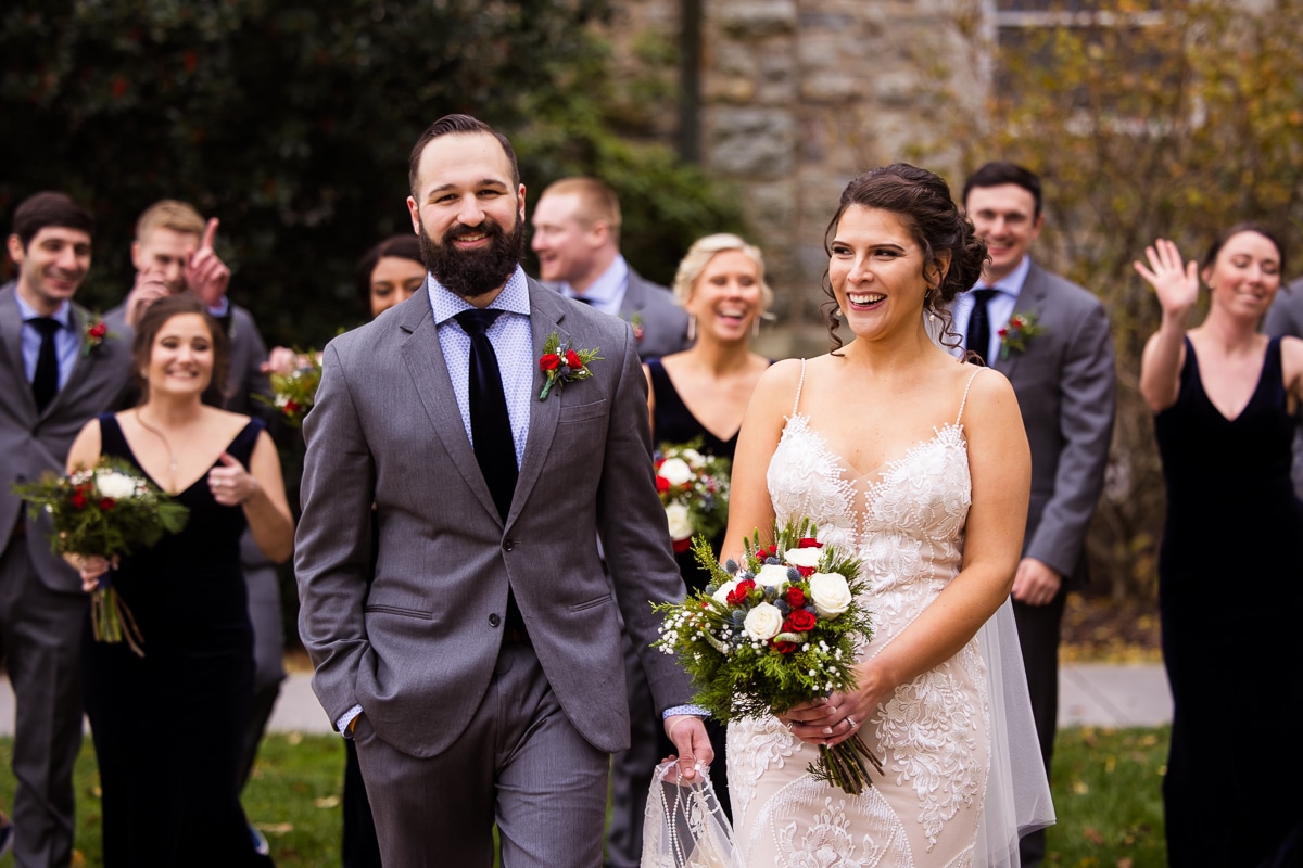 mount st mary's wedding photographer, rhinehart photography, captures the bride and groom walking and laughing as they wedding party cheers and dances behind them outside of the chapel before their wedding ceremony