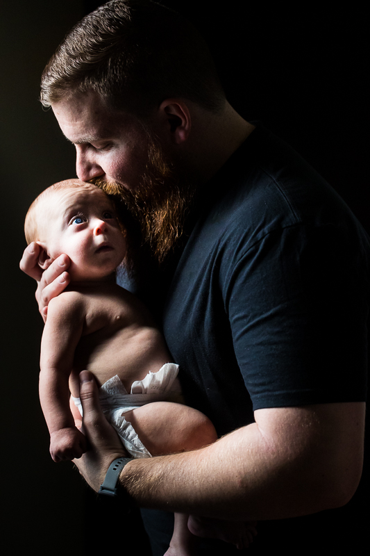 Lehigh Valley Family Photographer, rhinehart photography, captures dad as he kisses his son on the forehead with the baby's vibrant blue eyes looking up during this newborn family session 