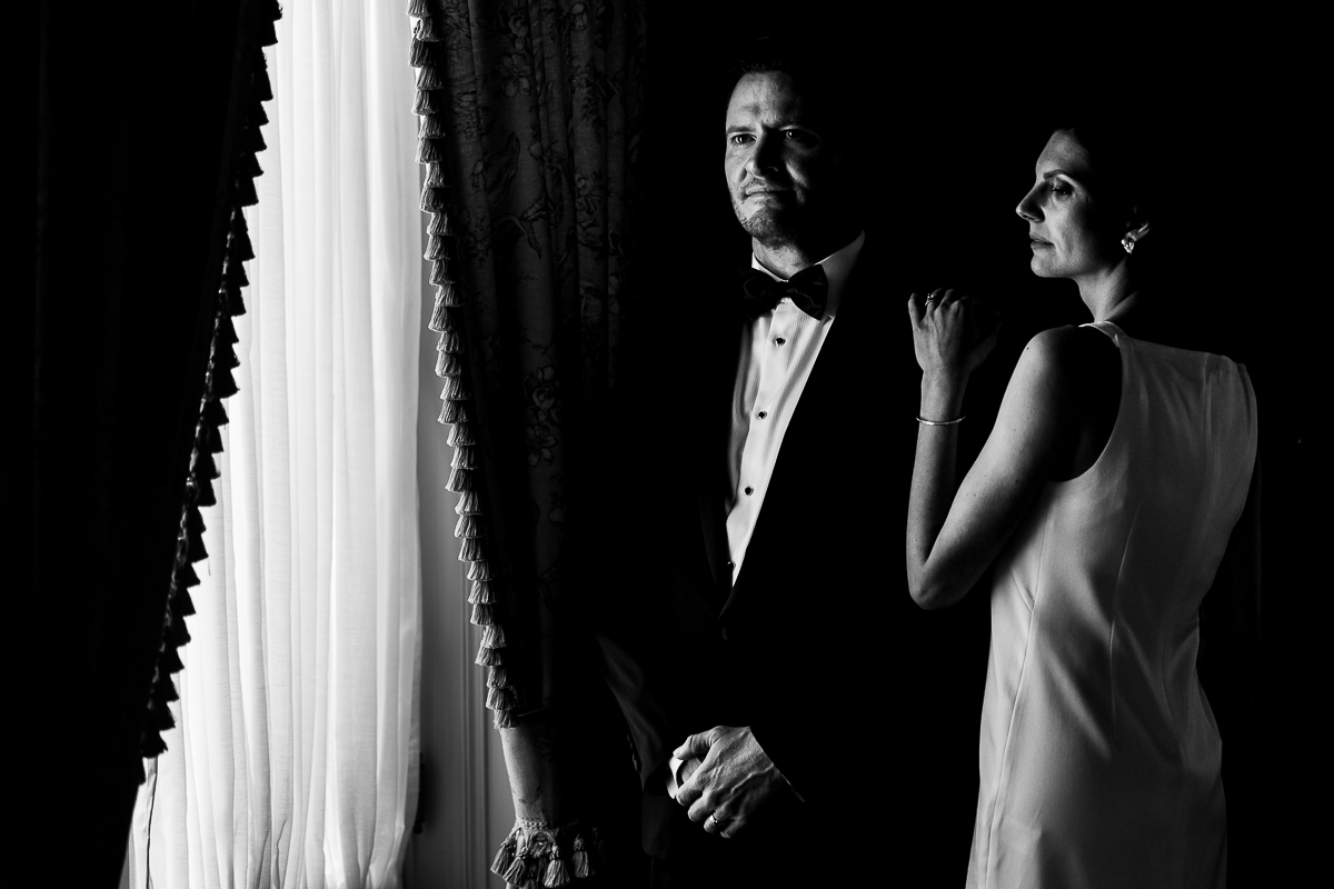 Timeless, elegant black and white image of the bride and groom standing together near a window as the light from the window reflects on their faces