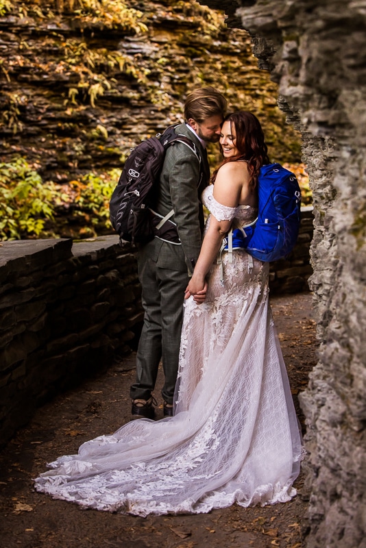 vibrant, colorful image of the couple holding hands as they are surrounded by rock formations and vibrant fall leaves in their wedding dress and suit with their hiking boots and backpack as they explore watkins glen state park together 