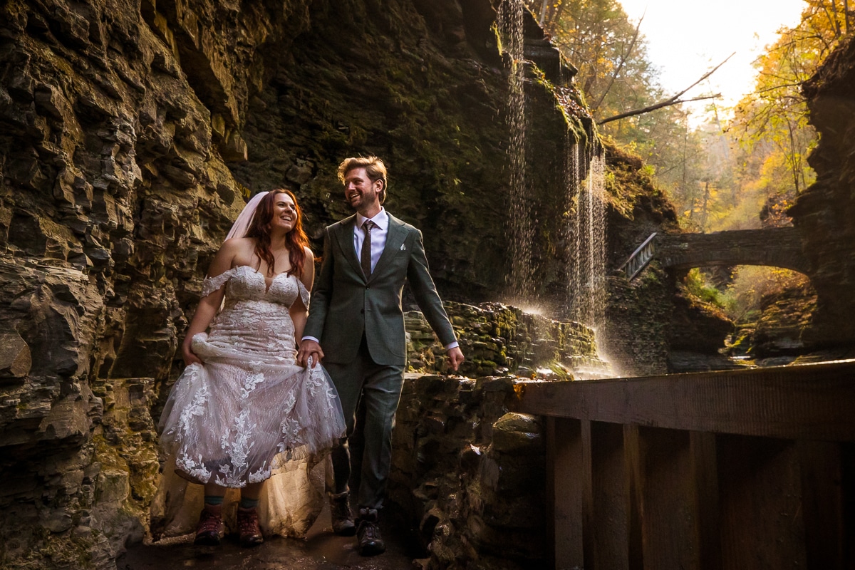 Watkins glen wedding photographer, rhinehart photography, captures this unique, authentic, creative image of the couple laughing and smiling at one another as they hike through watkins glen during their after session in their wedding attire as the sun beats down on them with a waterfall and rocks behind them