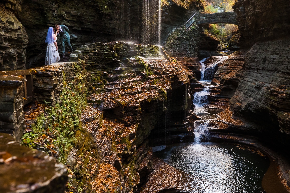 Watkins glen wedding photographer, rhinehart photography, captures this unique, adventurous image of the couple in their wedding attire exploring the state park with the water flowing beneath them and a waterfall behind them 