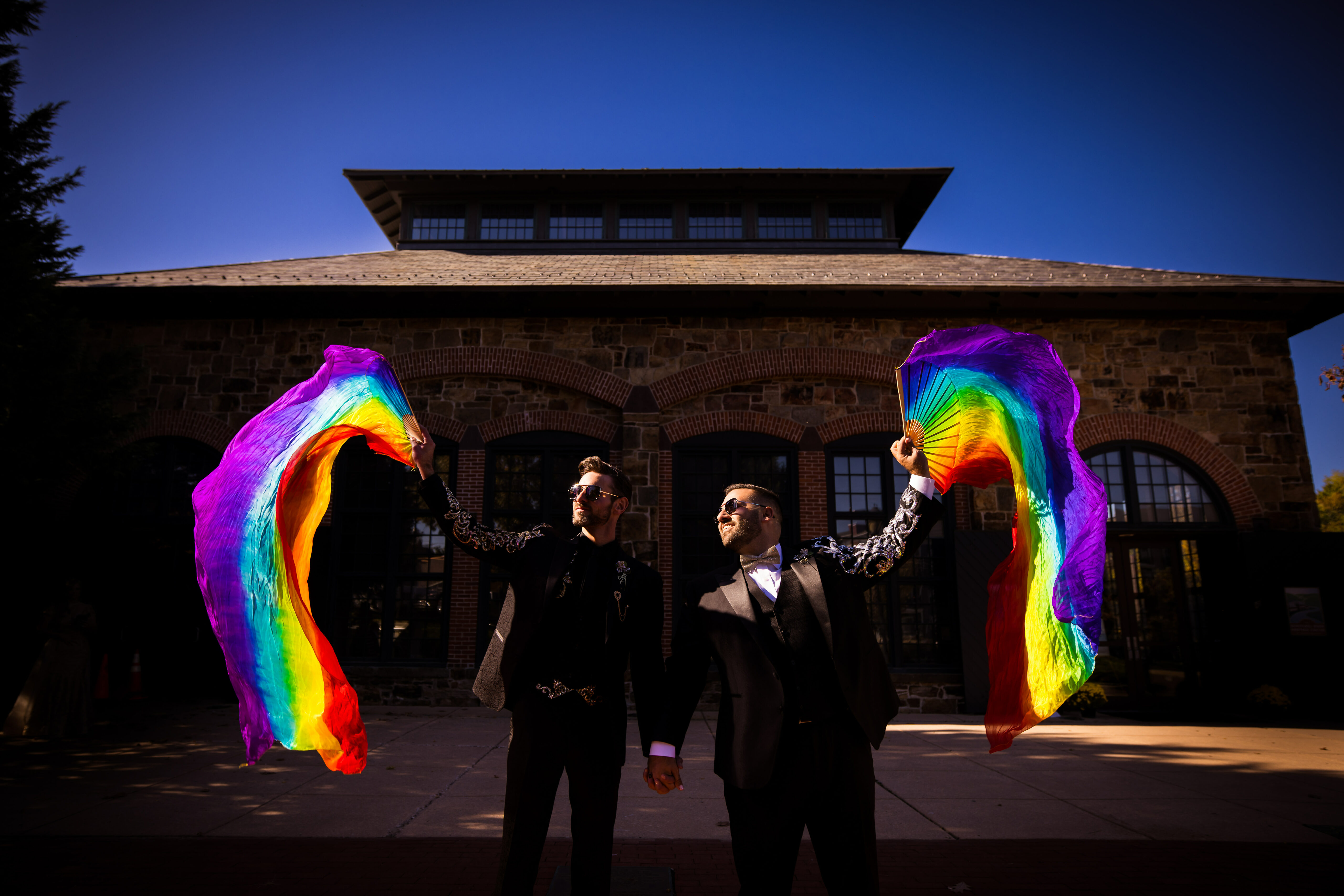 LGBTQ+ Wedding Photographer, Lisa Rhinehart, captures this colorful, fun image of the grooms holding hands as they fly rainbow fans in the air together at their Phoenixville Foundry wedding venue