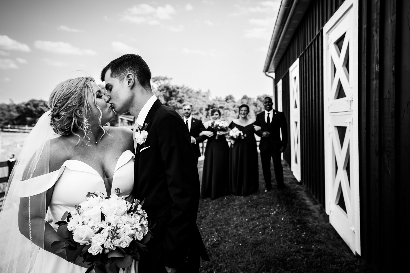 Middleburg Barn Wedding Photographer, lisa rhinehart, captures this black and white image of the couple kissing outside of the barn with their wedding party cheering and smiling behind them 