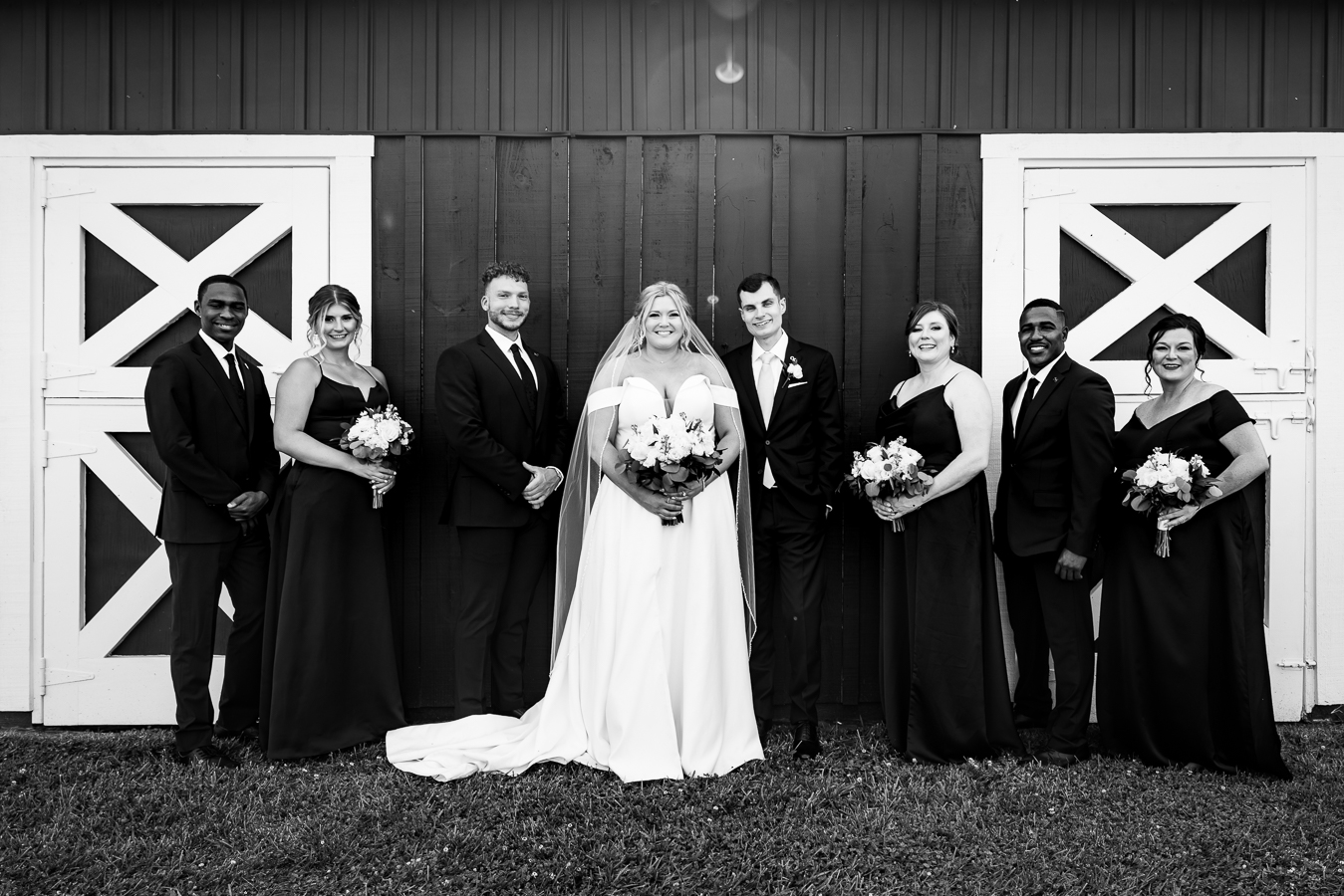 traditional black and white image of the bride and groom with their wedding party standing outside of the barn in a line 