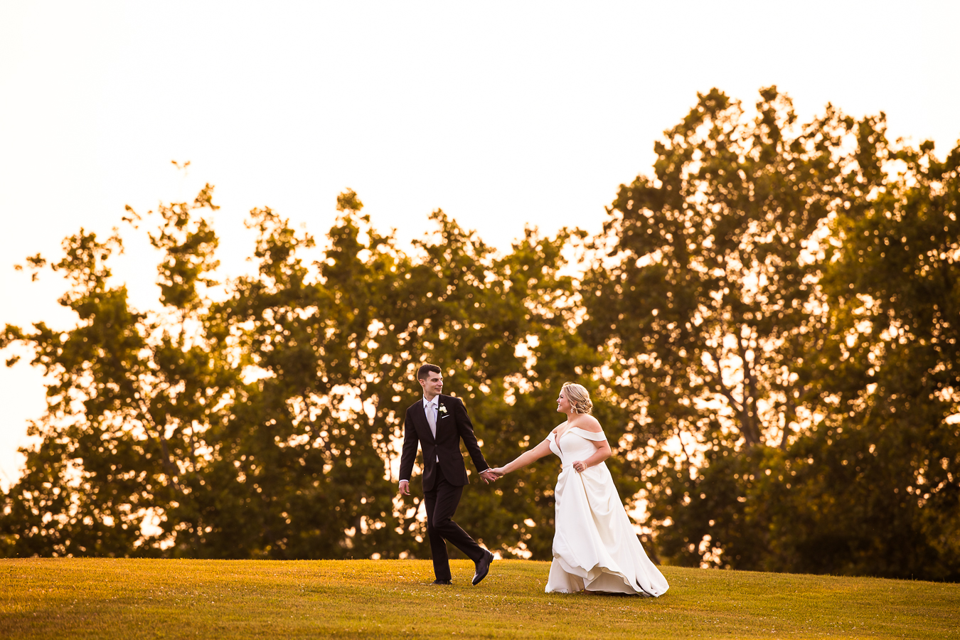 rhinehart photography captures this candid, creative image of the bride and groom as they run across and open field holding hands