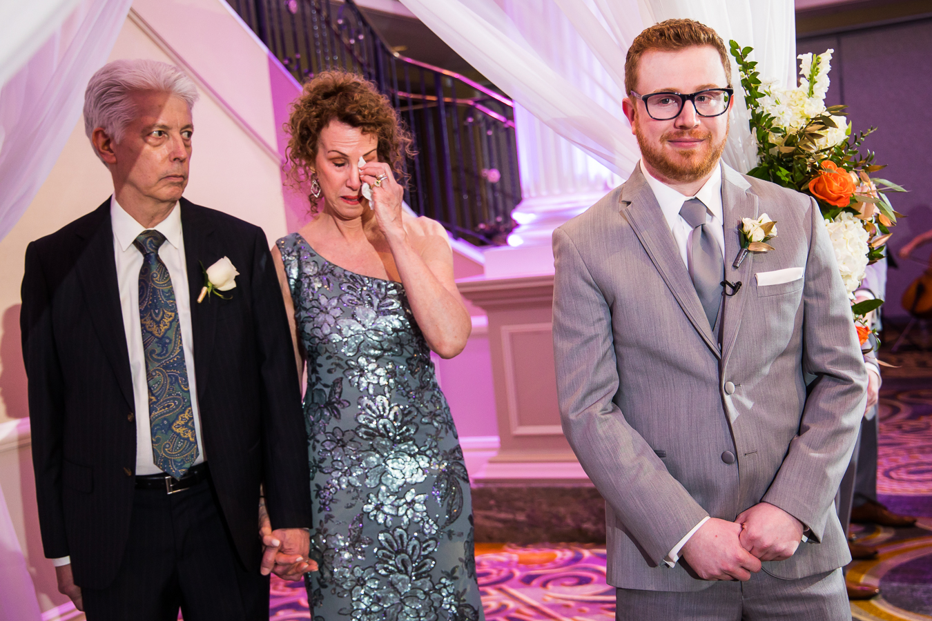 palace at somerset park photographer, lisa rhinehart, captures this authentic image of the groom looking at his bride as she walks down the aisle with his parents standing behind him wiping away tears during this palace at somerset wedding ceremony 