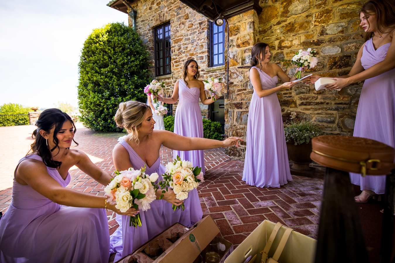 image of the bridesmaids in their lilac colored dress passing flower bouquets around while the bride gets pictures taken in the background behind them before the wedding ceremony 