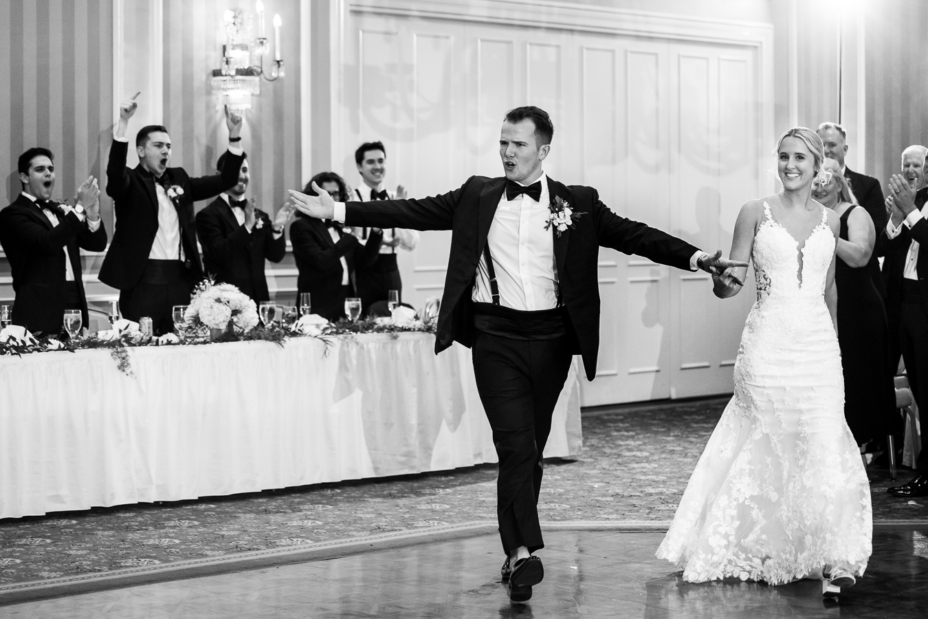 Country Club of York Wedding photographer, lisa rhinehart, captures this black and white image of the bride and groom as they walk into their wedding reception together as everyone is cheering and clapping around them